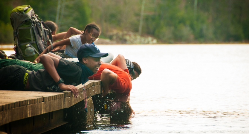 A group of students lay on a dock above a body of water. One of them dips the top of their head into the water. 